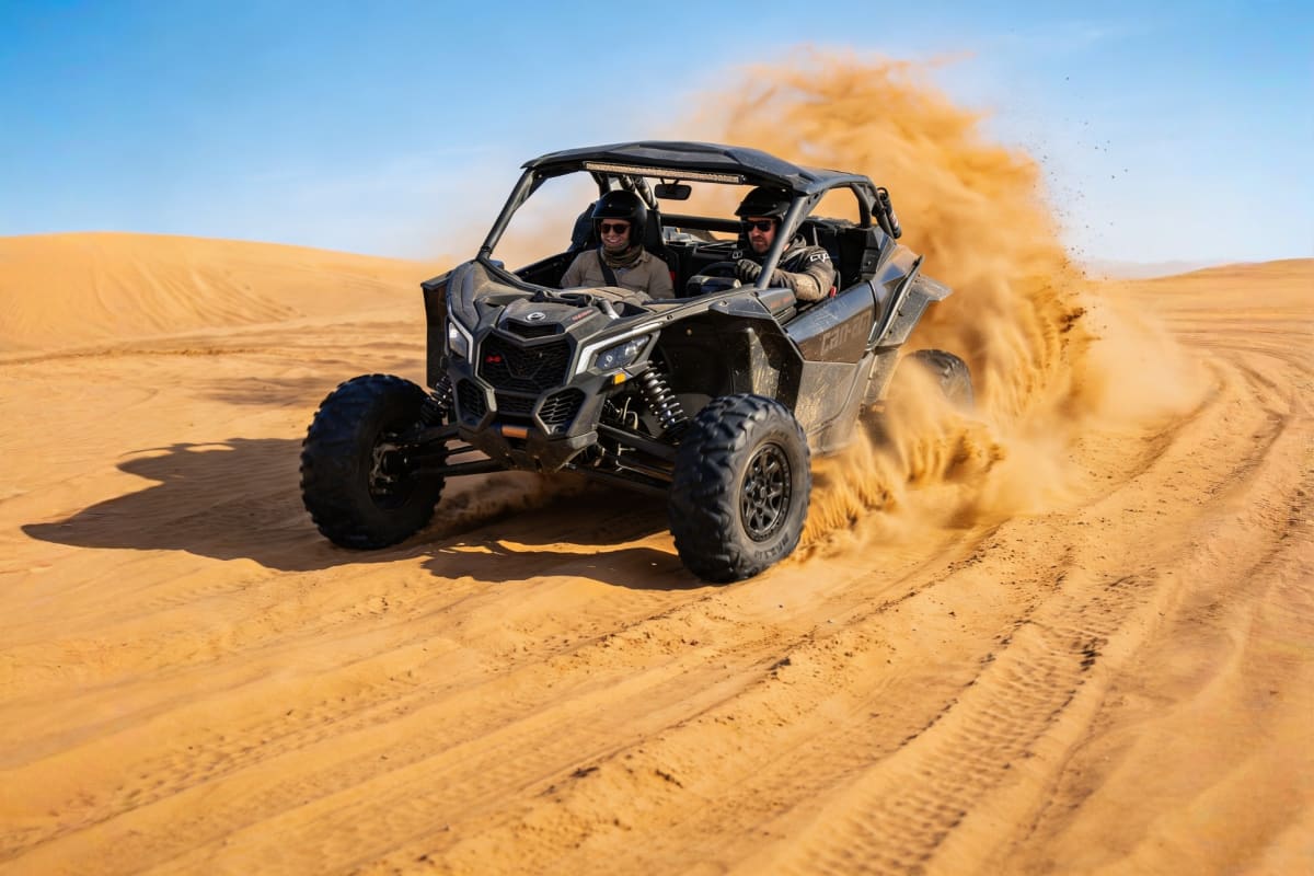 Two riders in a Can-Am Maverick buggy kicking up dust while driving across golden sand dunes in the desert
