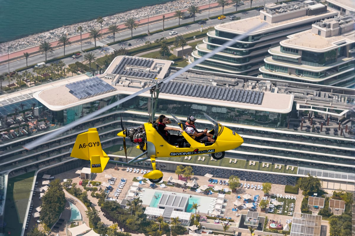 Yellow gyrocopter flying over Dubai cityscape with Burj Khalifa and modern skyscrapers visible below against blue sky (3)
