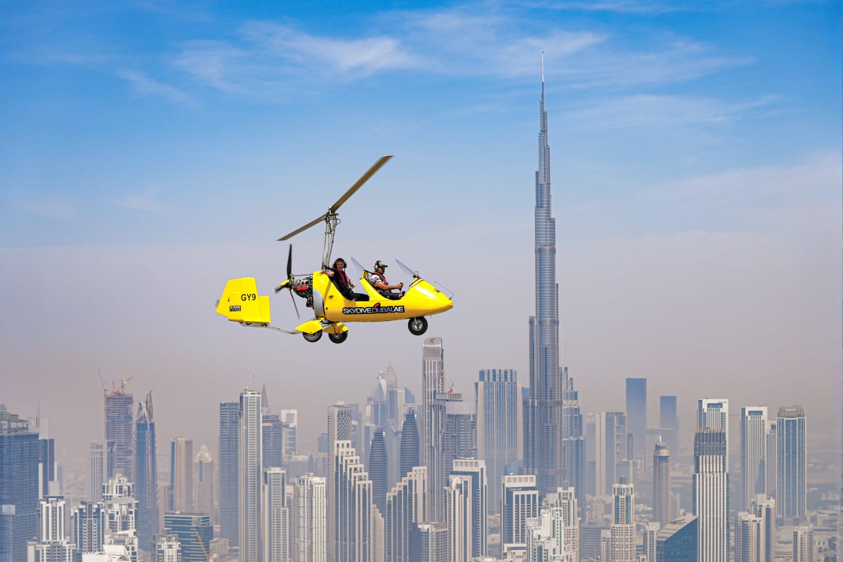 Yellow gyrocopter flying over Dubai cityscape with Burj Khalifa and modern skyscrapers visible below against blue sky