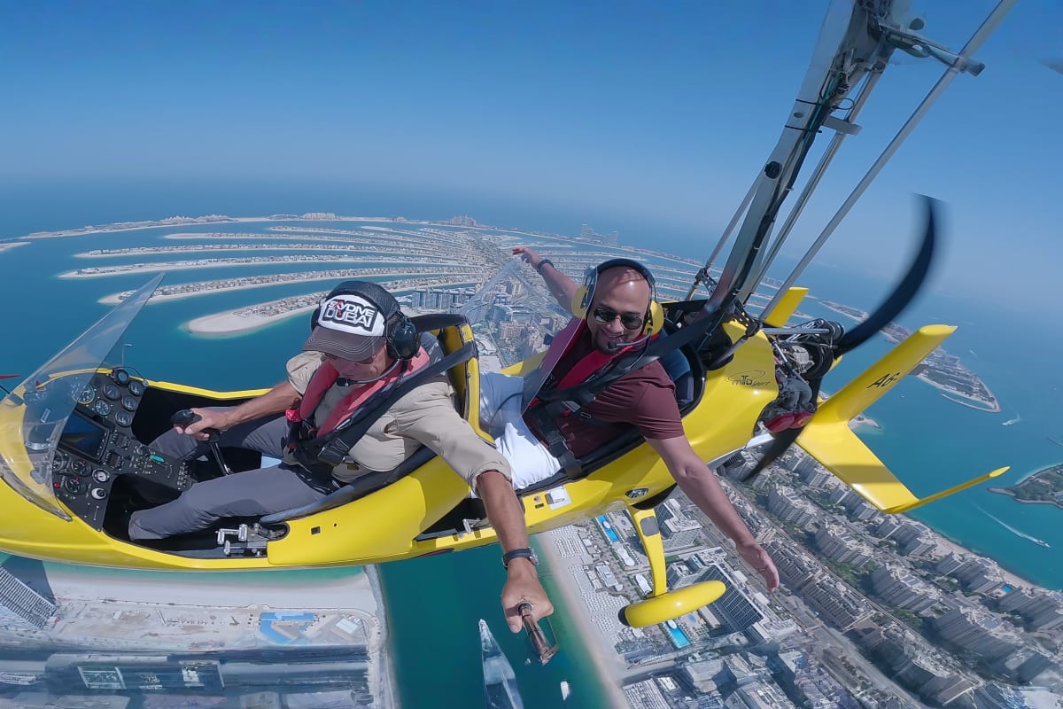 Yellow gyrocopter flying over Dubai cityscape with Burj Khalifa and modern skyscrapers visible below against blue sky (4)