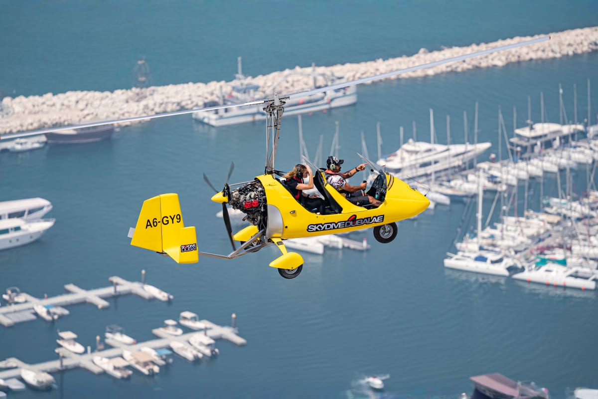 Yellow gyrocopter flying over Dubai cityscape with Burj Khalifa and modern skyscrapers visible below against blue sky (5)