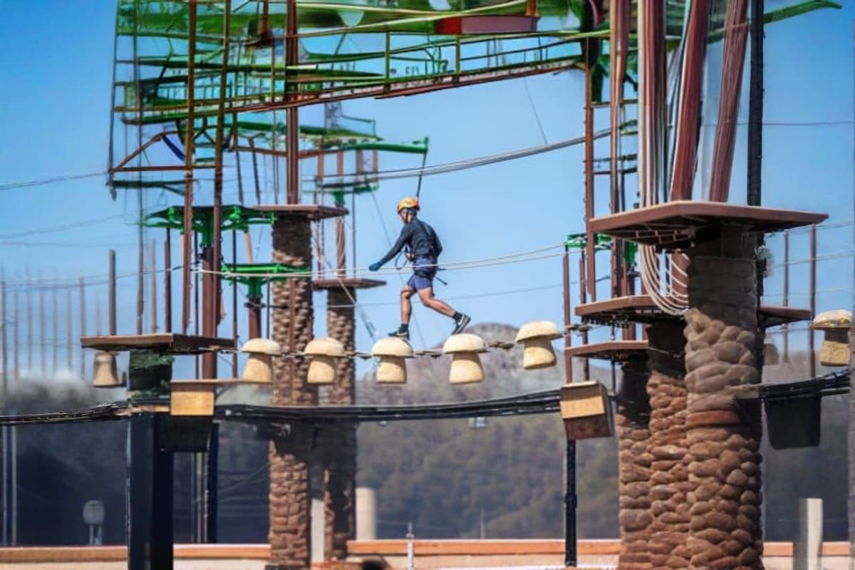 Person zip-lining across obstacle course with metal beams and stone towers at adventure park in mountainous landscape