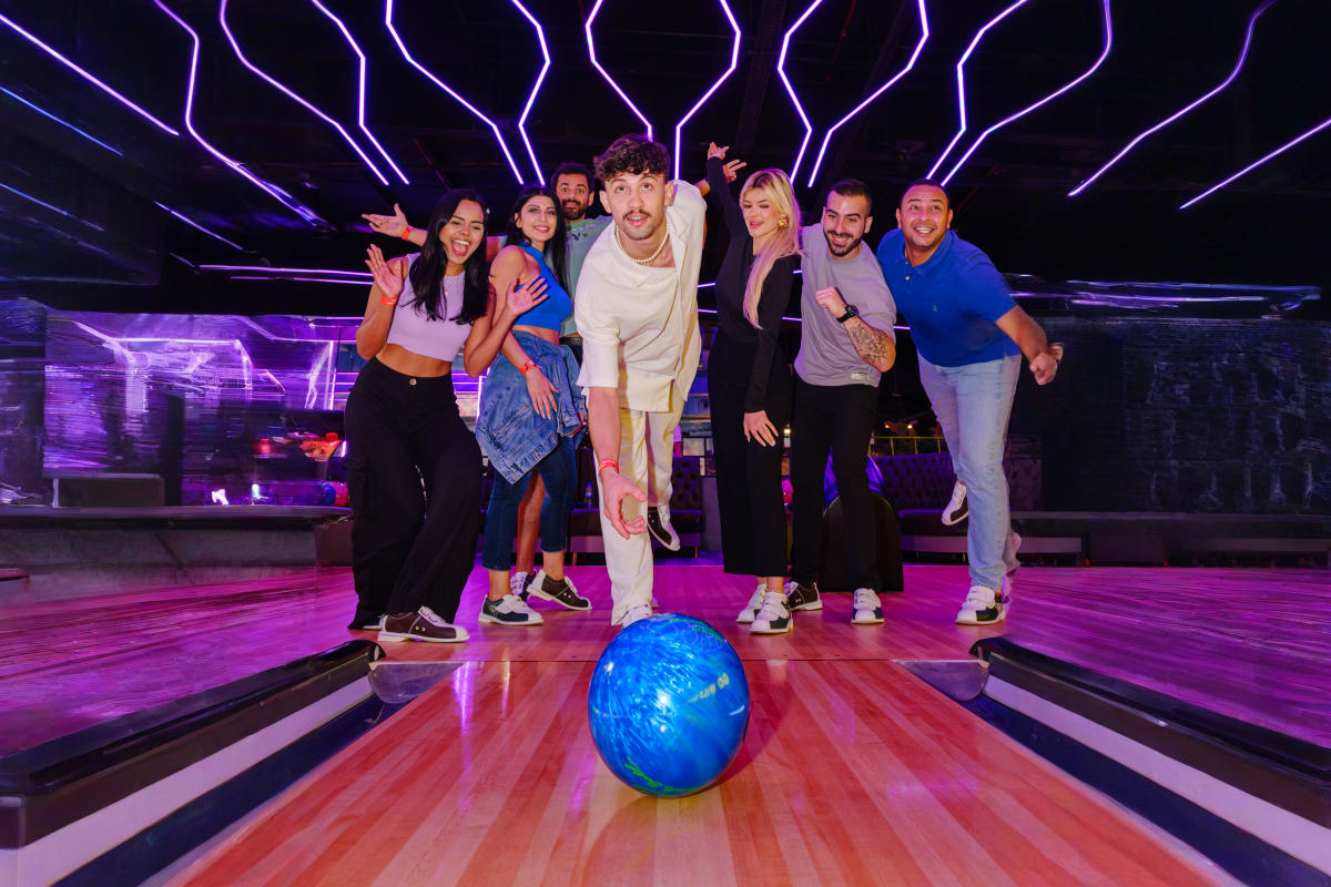 Group of eight people posing at a bowling alley with a blue bowling ball on the lane, purple and pink neon lighting